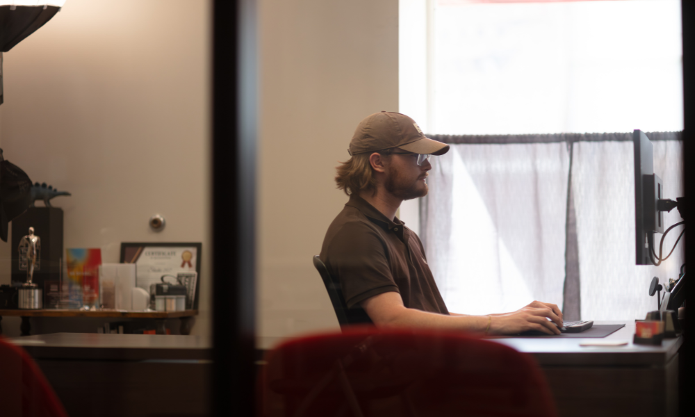 man wearing glasses and hat working at a computer at a desk in a glass office space