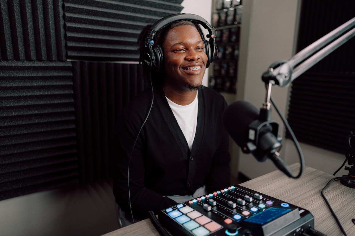 A student sitting inside of a recorsing podcast studio smiling. He has headphones on and a microphone in front of him.