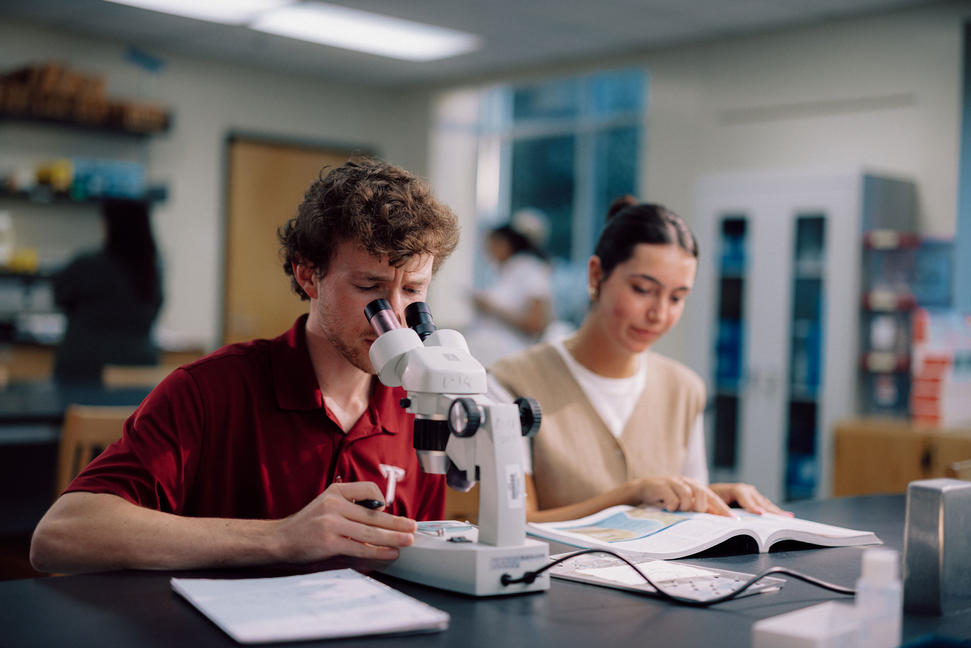 Two biology students observe a sample under a microscope.