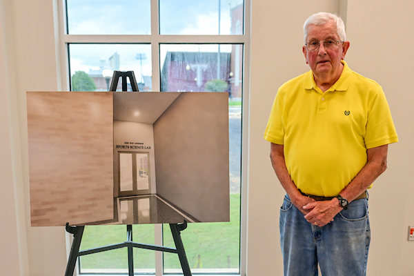 John "Doc" Anderson standing with the rendering of the new sports lab with his name above it. 